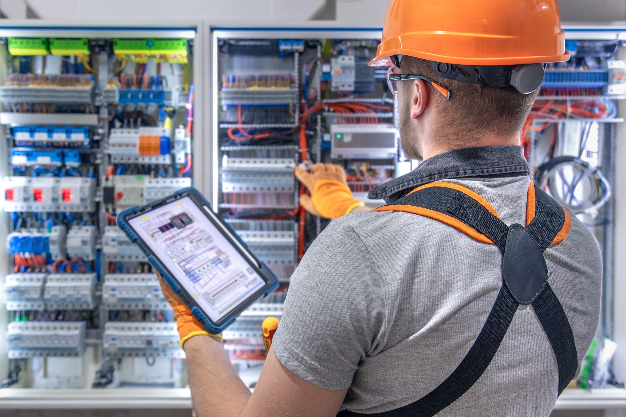 An electrician in a hard hat using a tablet in front of a complex electrical switchboard
