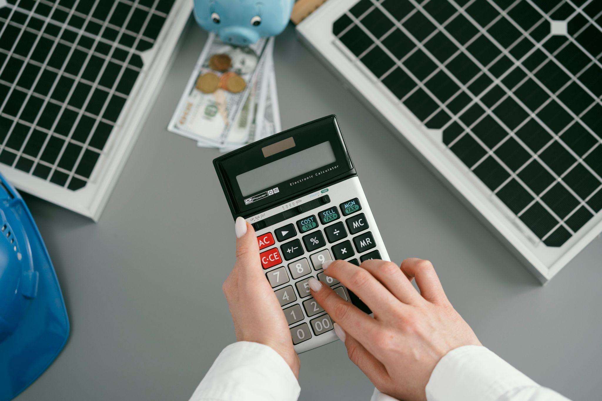 A person calculating costs on a calculator beside a solar panel, banknotes and a piggy bank