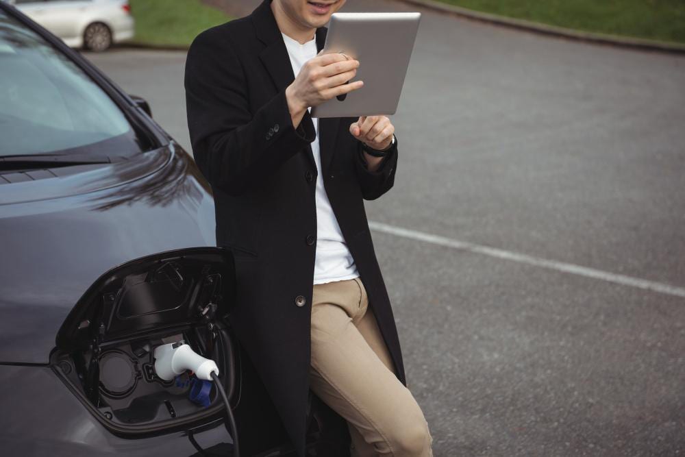 A person leaning on an electric car using a tablet while the car charges at the kerbside